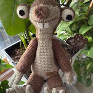 May include: A handmade crochet sloth doll in shades of brown and beige. The sloth has large, expressive eyes with black pupils, a prominent nose, and two large teeth. The doll is posed with its arms and legs slightly outstretched, and a small "handmade" tag is attached.