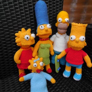 Può includere: Una famiglia di personaggi dei Simpson realizzata a uncinetto. La famiglia comprende Homer, Marge, Bart, Lisa e Maggie. I personaggi indossano tutti i loro abiti caratteristici.