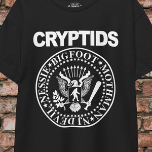 Könnte beinhalten: Schwarzes T-Shirt mit einer weißen Grafik eines Siegels mit den Worten "CRYPTIDS", "BIGFOOT", "MOTHMAN", "NESSIE" und "DEVIL" um das Siegel herum.