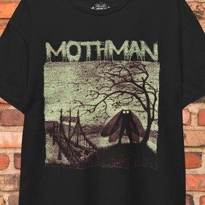 Könnte beinhalten: Schwarzes T-Shirt mit einem grafischen Aufdruck von Mothman, einem Kryptiden mit großen Augen und Flügeln, der auf einer Brücke in einem Wald steht. Der Text "Mothman" ist über dem Bild gedruckt.