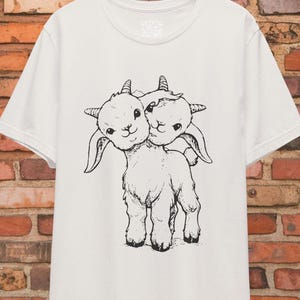 May include: White t-shirt featuring a black and white illustration of a two-headed goat.