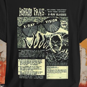 Può includere: Maglietta nera con una grafica in stile fumetto vintage che presenta un paio di occhiali a raggi X e il testo "Horror Fans: See Living "Skeletons" with these Mystery X-Ray Glasses". Il testo "Vision" e "Only $1.00" sono anche presenti sulla grafica.
