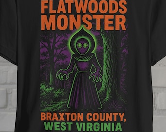 May include: Black t-shirt featuring a graphic of the Flatwoods Monster in a forest setting. The design includes the text "Flatwoods Monster" in orange and "Braxton County, West Virginia" in green.
