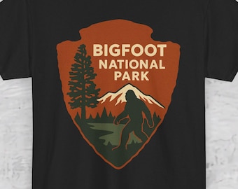 Pode incluir: Camiseta preta com um design do Parque Nacional Bigfoot. O gráfico inclui uma forma de ponta de flecha marrom com um pinheiro, montanhas e uma silhueta de Bigfoot. O texto "BIGFOOT NATIONAL PARK" é marrom claro.