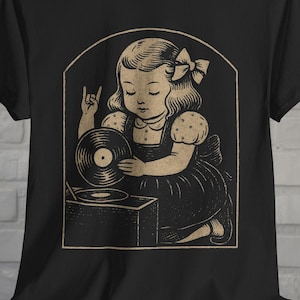May include: Black t-shirt with a sepia-toned vintage illustration. It depicts a girl with a bow, holding a vinyl record and giving the rock and roll hand sign. The design is framed within an arched shape.