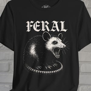 Puede incluir: Una camiseta negra de manga corta con una ilustraci&oacute;n gr&aacute;fica blanca de una zarig&uuml;eya siseando. Sobre la zarig&uuml;eya, la palabra FERAL est&aacute; impresa en una fuente blanca, negrita y estilizada, centrada en la parte frontal de la prenda.