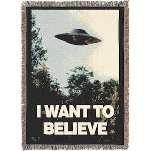 May include: Woven blanket with a black background featuring a UFO flying over trees. The text "I WANT TO BELIEVE" is displayed in white block letters. The blanket has a fringed edge.