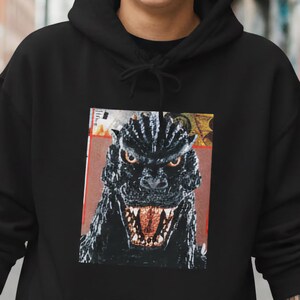 May include: Black hoodie with a Godzilla graphic print. The image shows the monster with an open mouth and sharp teeth. The graphic is set against a red and white background. A comfortable, casual garment.