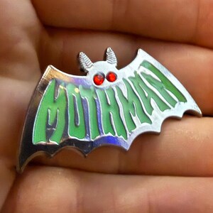 May include: A silver-colored Mothman pin in the shape of a bat. The pin has green and silver wings with the word "MOTHMAN" in green lettering. The eyes are red, and the pin is held in a hand.
