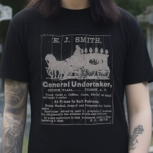 May include: A black t-shirt with a vintage advertisement for "E. J. Smith, General Undertaker" printed on the front. The advertisement features a horse-drawn hearse and text that reads "Church Plaza, Tucson, A. T."