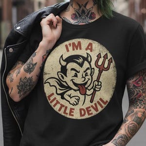 May include: Black t-shirt featuring a vintage-style graphic of a cartoon devil holding a pitchfork, with the text "I'M A LITTLE DEVIL" in a circular design. The design is in shades of red, beige, and black.