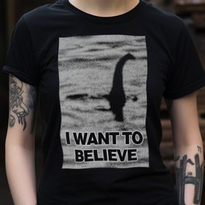 Model wearing a Loch Ness Monster believer T-shirt, featuring iconic "I Want to Believe" phrase with Nessie silhouette against a scenic lake background, perfect for fans of cryptozoology and unexplained mysteries.