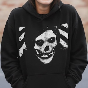 May include: Black hooded sweatshirt featuring a white skull graphic with a distressed design. The sweatshirt has a front pocket and a drawstring hood. The skull graphic is centered on the front of the sweatshirt.