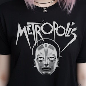 May include: A woman with pink hair wearing a black t-shirt with a white graphic of a face and the word "Metropolis".