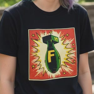 Pode incluir: Camiseta preta com um gr&aacute;fico de uma bomba com a letra "F" dentro de uma explos&atilde;o amarela, emoldurada por uma borda vermelha com estrelas. A pessoa que usa a camiseta tem cabelo multicolorido.