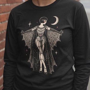 Könnte beinhalten: Ein schwarzes Langarmshirt mit einer grafischen Illustration einer Figur mit Bob-Frisur, die einen Umhang mit Spinnennetz-Muster trägt. Das Design zeigt einen Halbmond und kleine Sterne in den Farben Creme, Schwarz und zartem Rosa auf der Brust.