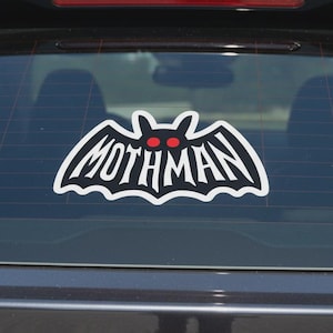 May include: A white vinyl sticker featuring the word "MOTHMAN" in a stylized font, set within a bat-like silhouette. The bat has red eyes. The sticker is affixed to the rear window of a vehicle.