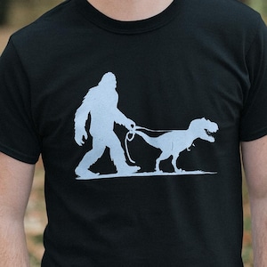 May include: Black t-shirt with a light blue graphic depicting Bigfoot walking a T-Rex on a lead. The design is a humorous take on pet ownership. The person wearing the shirt is smiling.