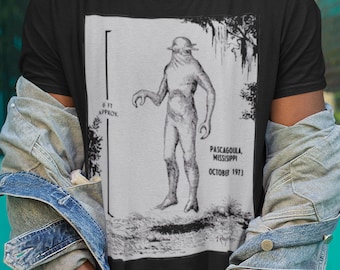 May include: Black t-shirt featuring a grayscale illustration of a humanoid figure with the text "Pascagoula Mississippi October 1973." The figure is depicted with a hat and claw-like hands. The shirt is paired with dark jeans and a denim jacket.