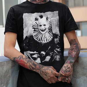 May include: A person wearing a black t-shirt with a black and white image of a clown. The t-shirt has a distressed look. The person has a nose ring and tattoos on their neck and arms. They are wearing black jeans with rips in the knees.
