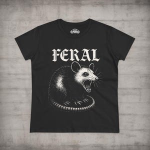Puede incluir: Una camiseta negra de manga corta con un estampado gr&aacute;fico blanco de una zarig&uuml;eya gru&ntilde;endo. La palabra FERAL est&aacute; impresa en una fuente blanca, negrita y con serifa sobre la ilustraci&oacute;n de la zarig&uuml;eya. Tiene cuello redondo y un logo interior.