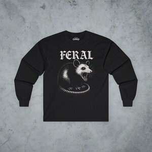 Puede incluir: Una camiseta negra de manga larga con una ilustraci&oacute;n gr&aacute;fica blanca de una zarig&uuml;eya siseando centrada en el pecho. Sobre la zarig&uuml;eya, la palabra FERAL est&aacute; impresa en una fuente g&oacute;tica blanca y negrita. La camiseta tiene cuello redondo.