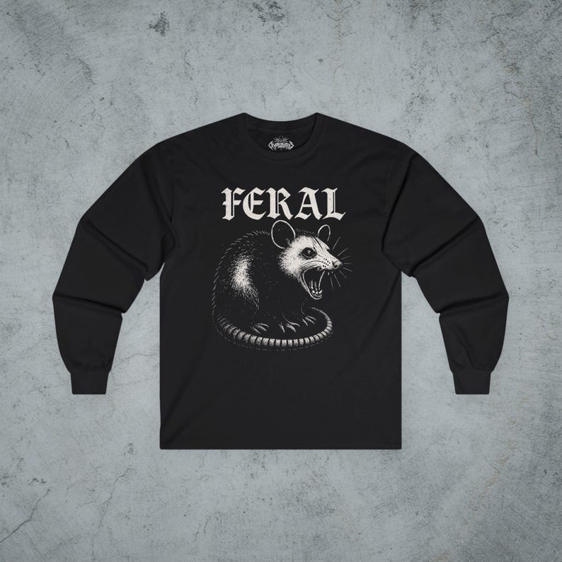 Puede incluir: Una camiseta negra de manga larga con una ilustraci&oacute;n gr&aacute;fica blanca de una zarig&uuml;eya siseando centrada en el pecho. Sobre la zarig&uuml;eya, la palabra FERAL est&aacute; impresa en una fuente g&oacute;tica blanca y negrita. La camiseta tiene cuello redondo.