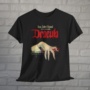May include: A black short-sleeve t-shirt featuring a graphic print of a pale hand with long fingernails resting on a dark surface. Above the hand, the text reads Bram Stoker's Original Dracula in red and white lettering against the black fabric.
