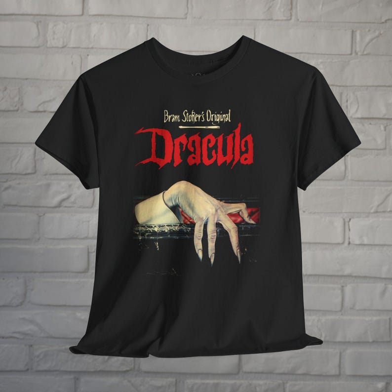 May include: A black short-sleeve t-shirt featuring a graphic print of a pale hand with long fingernails resting on a dark surface. Above the hand, the text reads Bram Stoker's Original Dracula in red and white lettering against the black fabric.