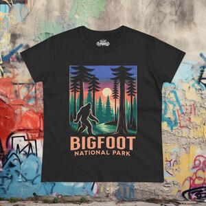 May include: Black t-shirt featuring a Bigfoot graphic in a forest scene with a sunset. The design includes the text "BIGFOOT NATIONAL PARK" in a peach color. The shirt has a crew neck and short sleeves.