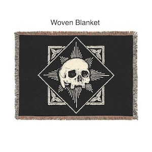 May include: Black woven blanket featuring a white skull design within a diamond-shaped frame. The frame is surrounded by a radiating pattern and floral accents. The blanket has a fringed edge and the text "Woven Blanket" is at the top.