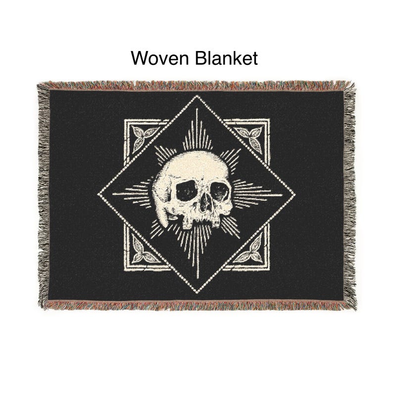 May include: Black woven blanket featuring a white skull design within a diamond-shaped frame. The frame is surrounded by a radiating pattern and floral accents. The blanket has a fringed edge and the text "Woven Blanket" is at the top.