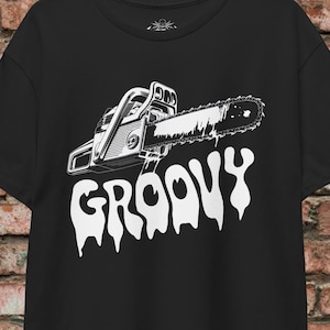 Puede incluir: Camiseta negra con un gráfico blanco de una motosierra y la palabra "GROOVY" en una fuente goteante. El diseño está centrado en la parte delantera. La camiseta tiene mangas cortas y cuello redondo.
