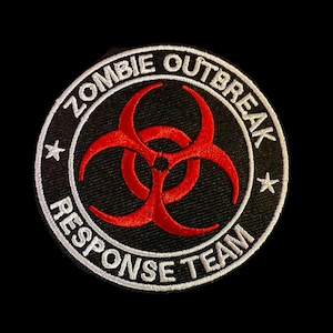 May include: Embroidered patch with the text "ZOMBIE OUTBREAK RESPONSE TEAM" in white lettering on a black background. The patch features a red biohazard symbol in the center and two white stars. The patch is circular in shape.