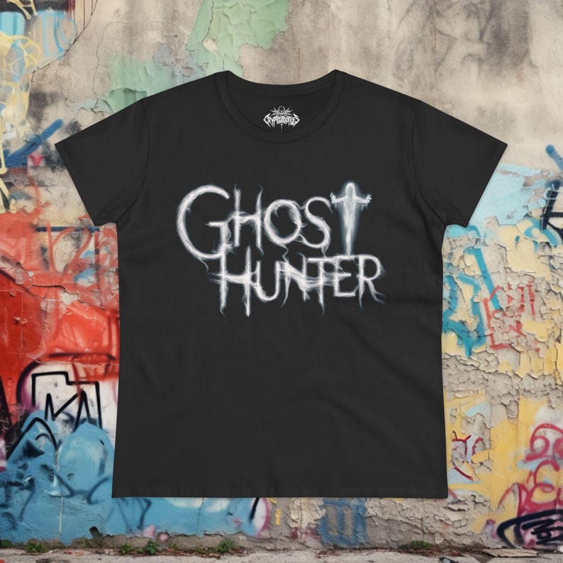 May include: A black short-sleeve t-shirt featuring the white text Ghost Hunter in a wispy, ethereal font style across the chest. The letter T in Ghost is stylized as a ghostly figure with arms raised. A small logo is printed inside the neck area.