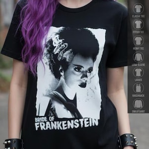 Pode incluir: Uma t-shirt preta com uma estampa gráfica branca da Noiva de Frankenstein com o seu penteado característico. O texto "BRIDE OF FRANKENSTEIN" está impresso a branco por baixo do retrato. A t-shirt tem mangas curtas e gola redonda.