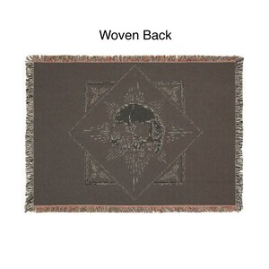 May include: A dark brown woven throw blanket with a skull design in the center. The skull is surrounded by a starburst pattern and geometric shapes. The blanket has a fringed edge and the text "Woven Back" at the top.