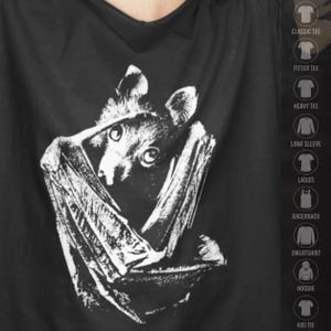 Puede incluir: Una camiseta negra con una ilustración gráfica blanca de un murciélago con las alas envueltas alrededor de su cuerpo. En el lado derecho, una lista vertical de opciones de prendas incluye los textos: CLASSIC TEE, FITTED TEE, HEAVY TEE, LONG SLEEVE, LADIES, RACERBACK, SWEATSHIRT, HOODIE y KIDS TEE.