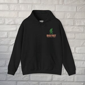May include: Black hooded sweatshirt with a front pocket and drawstring. The design features a green silhouette of Bigfoot above the words "BIGFOOT NATIONAL PARK" in orange and white.