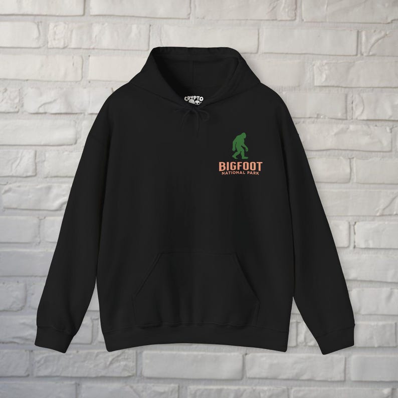 May include: Black hooded sweatshirt with a front pocket and drawstring. The design features a green silhouette of Bigfoot above the words "BIGFOOT NATIONAL PARK" in orange and white.