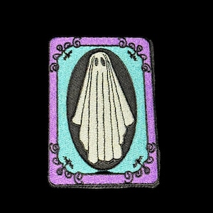 May include: Embroidered patch featuring a white ghost with a simple face inside a black oval. The oval is set against a light blue background, framed by a purple border with decorative swirls. A spooky Halloween-themed design.