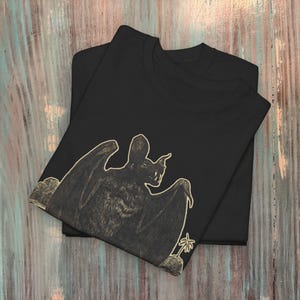 May include: Black t-shirt with a graphic print of a bat in a vintage style. The bat is outlined in a light beige color and is perched on a small rock with a palm tree in the background.