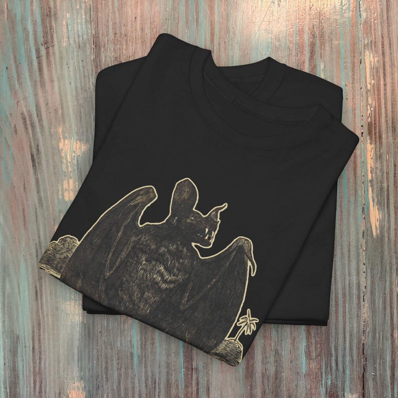 May include: Black t-shirt with a graphic print of a bat in a vintage style. The bat is outlined in a light beige color and is perched on a small rock with a palm tree in the background.