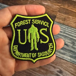 May include: A green and black embroidered patch with the text "Forest Service" and "U.S. Department of Sasquatch" surrounding a green silhouette of a sasquatch.
