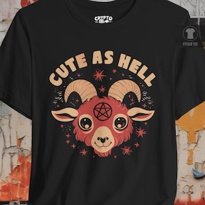 May include: A black t-shirt featuring a graphic of a red goat head with large eyes, curved horns, and a black pentagram on its forehead. The text "CUTE AS HELL" is printed in a curved beige font above the goat, surrounded by small red stars.