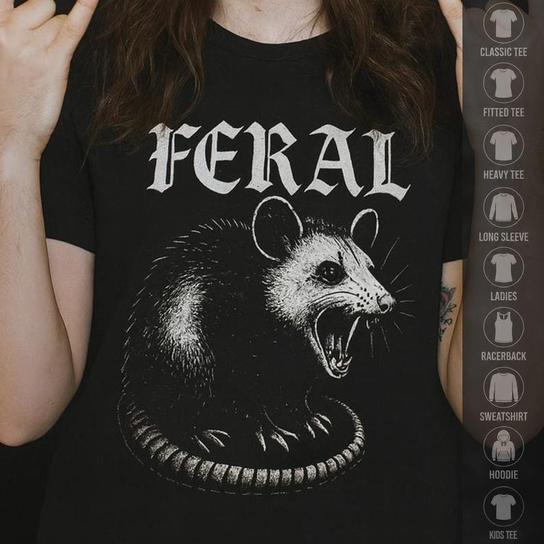 Puede incluir: Una camiseta negra de manga corta con una ilustraci&oacute;n gr&aacute;fica blanca de una zarig&uuml;eya siseando y la palabra FERAL impresa en una fuente g&oacute;tica negrita y desgastada encima del animal. El dise&ntilde;o est&aacute; centrado en el pecho de la prenda.