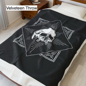 May include: Black velveeteen throw blanket with a white skull design. The skull is centered within a white diamond shape, surrounded by radiating lines and decorative corner elements. The blanket is on a bed with a white pillow.