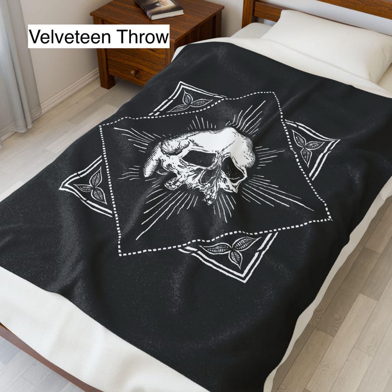 May include: Black velveeteen throw blanket with a white skull design. The skull is centered within a white diamond shape, surrounded by radiating lines and decorative corner elements. The blanket is on a bed with a white pillow.