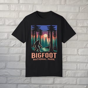 May include: Black t-shirt featuring a Bigfoot graphic in a forest scene with a sunset backdrop. The design includes the text "BIGFOOT NATIONAL PARK" in a bold, orange font. The graphic depicts a silhouette of Bigfoot walking.