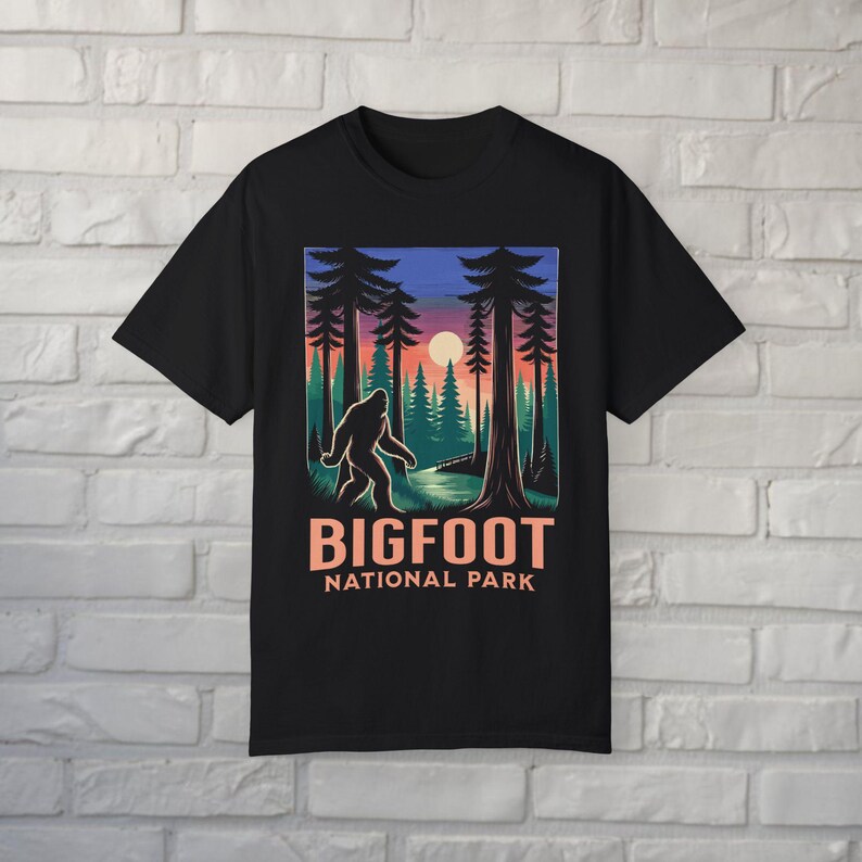 May include: Black t-shirt featuring a Bigfoot graphic in a forest scene with a sunset backdrop. The design includes the text "BIGFOOT NATIONAL PARK" in a bold, orange font. The graphic depicts a silhouette of Bigfoot walking.